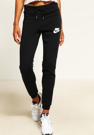 Persoon draagt zwarte Nike joggingbroek met wit logo en witte Nike sneakers met zwarte accenten, staande op een lichte vloer en achtergrond.