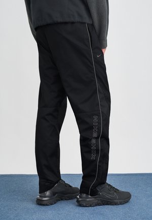 Personne portant un pantalon de survêtement Nike Shox noir et des baskets Nike noires debout sur un tapis bleu avec un fond clair uni.