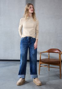 Beżowy top z długim rękawem, niebieskie szerokie dżinsy z podwiniętymi nogawkami oraz beżowe zamszowe buty na platformie, na tle betonowej ściany z drewnianym krzesłem.