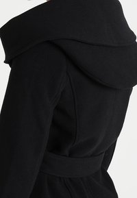 Schwarzer Mantel mit einer großen, gefalteten Kapuze, tailliertem Design und strukturiertem Stoff. Das Design ist figurbetont mit einer eleganten Silhouette.