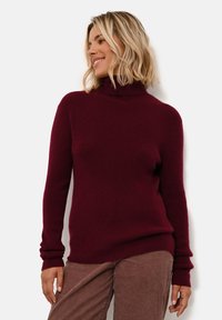 Burgundský rolák z pleteného materiálu, s dlouhými rukávy a žebrovanými manžetami, v uvolněném střihu. Nosený s hnědými kordověými kalhotami.