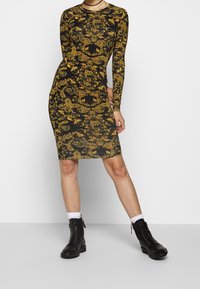 Långärmad, figurnära klänning i svart tyg med intrikata guld paisley- och blommönster. Har en midjelång fåll och rund halsringning.