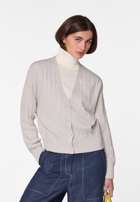 Helle beige, gerippte Strickjacke mit V-Ausschnitt, fünf Knöpfen und einem taillierten Saum. Getragen über einem weißen Rollkragenpullover und einer dunkelblauen, weiten Hose.