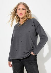 Sudadera gris con bordado de estrella negra, escote redondo, mangas largas y detalles de dobladillo sin terminar. Combinada con jeans oscuros.