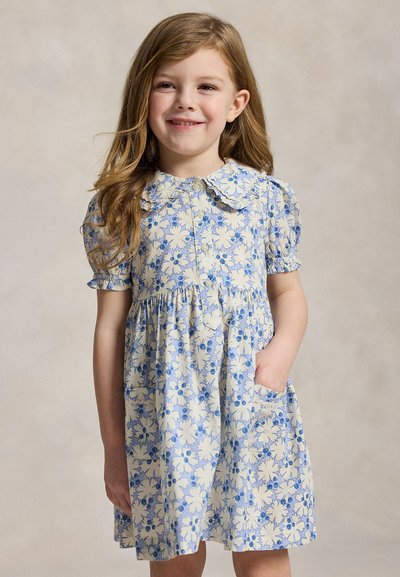 Ung jente smiler, iført en kortermet blå og kremfarget blomsterkjole med peter pan-krage, hånden i lommen, mot ensfarget bakgrunn.