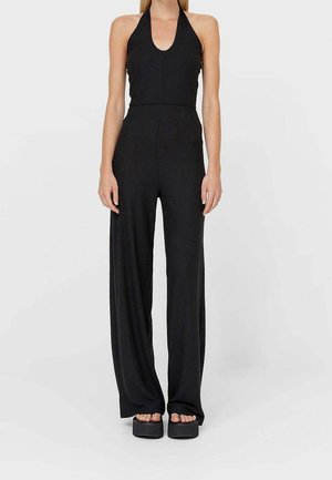 Black halter jumpsuit with a fitted bodice, wide-leg trousers, smooth texture, and deep neckline, paired with chunky black platform sandals.