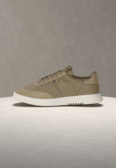 Μπεζ αθλητικό παπούτσι χαμηλού προφίλ με σχέδιο δέσιμου κορδονιών, λευκή σόλα και διακριτική λεπτομέρεια λογοτύπου στο πλάι, τοποθετημένο σε ουδέτερο φόντο.