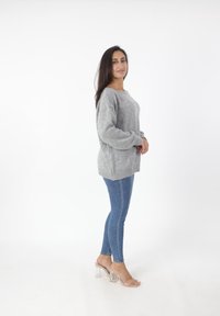 Maglione over-size grigio chiaro con orlo a coste, abbinato a jeans blu aderenti e sandali con tacco a blocco trasparente. Il modello è in profilo.