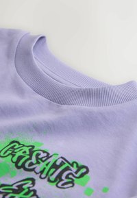 Vêtement à col ras du cou de couleur violet clair avec un col côtelé et un texte vert de style graffiti partiellement visible sur le tissu.