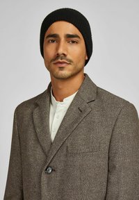 Bruine tweed blazer met een ingesneden revers, enkele knoopsluiting en een gestructureerde stof. Gecombineerd met een wit henley-shirt en een zwarte beanie.