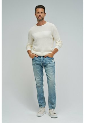 Hombre con suéter blanco de manga larga, vaqueros desgastados de color azul claro y zapatillas blancas, de pie con las manos en los bolsillos contra un fondo liso.