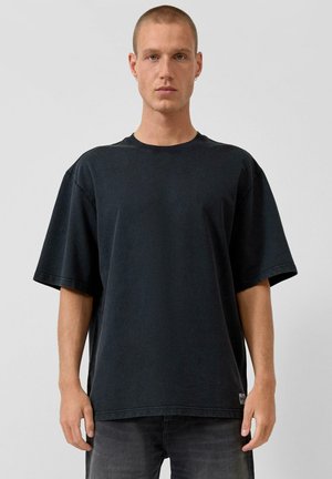 Młody mężczyzna z krótkimi włosami, ubrany w zwykły, luźny czarny T-shirt i ciemne dżinsy, stojący na jednolitym jasnym tle.