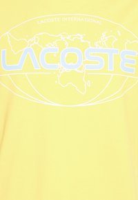 Žluté tričko s bílou grafikou světové mapy, nad níž je text "LACOSTE INTERNATIONAL" a pod ní "LACOSTE" v světle modré barvě.