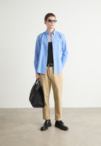 Lichtblauw overhemd met knopen, zwarte tanktop, beige broek, zwarte riem, zwarte schoenen en een zwart gemustreerde toilettas. Koele zonnebril accentueert de look.