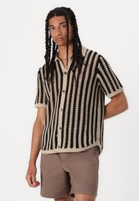 Maglia corta all'uncinetto a maniche corte a righe beige e nere, con colletto, chiusura con bottoni e motivo testurizzato. Abbinata a pantaloni corti marroni.