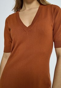 T-shirt en tricot à manches courtes en couleur rouille avec col en V. Présente une texture fine et douce avec des accents côtelés autour de l'encolure et des manches.
