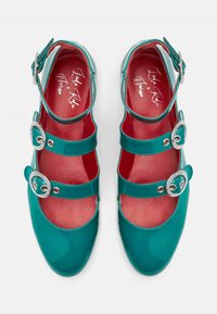Zapatos Mary Jane de charol color verde azulado con tres correas ajustables que presentan hebillas plateadas. El interior es de color rojo con una textura suave.