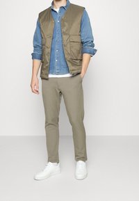 Gilet imbottito verde oliva sopra una camicia di jeans azzurra chiara, abbinato a pantaloni beige a sigaretta e sneakers bianche. Caratterizzato da tasche frontali e bottoni.