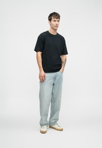 Μαύρο βαμβακερό t-shirt με κοντά μανίκια, συνδυασμένο με ανοιχτό μπλε τζιν. Το μοντέλο φορά λευκά αθλητικά παπούτσια με καφέ σόλες.