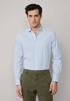 Chemise boutonnée rayée bleu clair et blanche en coton, avec un col pointu et une poche poitrine unique, portée avec un pantalon vert olive.