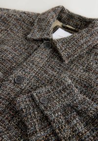 Chaqueta de camisa de cuadros oscuros hecha de tejido texturizado, con cuello y botones negros. Los colores incluyen gris, marrón y motas de crema.