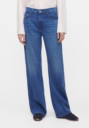 Personne portant un jean bleu à jambes larges, une chemise claire à rayures et des baskets foncées, debout devant un fond blanc uni.