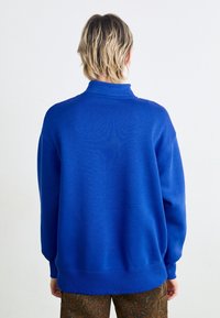 Modrý pulover mikina s vysokým límcem, volným střihem a žebrovanými manžetami. Hladká textura, jednobarevná a s minimálními designovými detaily.