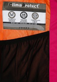 Etichetta in tessuto Clima Protect su materiale arancione e nero, che descrive caratteristiche impermeabili, traspiranti e resistenti al vento in più lingue.