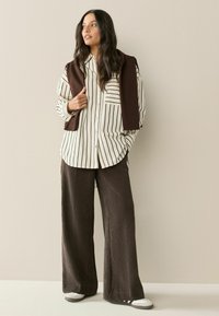 Gestreiftes weißes und schwarzes Hemd mit Knöpfen und Brusttasche, kombiniert mit braunen weiten, strukturierten Hosen und weißen Sneakers.