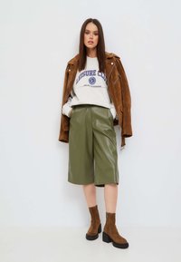 Brązowa skórzana kurtka z zamszu, szary sweatshirt z napisem "LEISURE CLUB", zielone skórzane szorty i brązowe botki na kostkę z grubym obcasem.