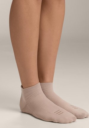 Lábak világos bézs bokazokniban, amelynek a lábujjaknál tapadós, texturált részletei vannak, semleges, egyszínű háttér előtt állva.