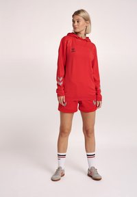 Röd idrottströja med huva, prickig design på axlarna och zigzag-accenter på ärmarna. Matchad med röda shorts och grå sneakers.
