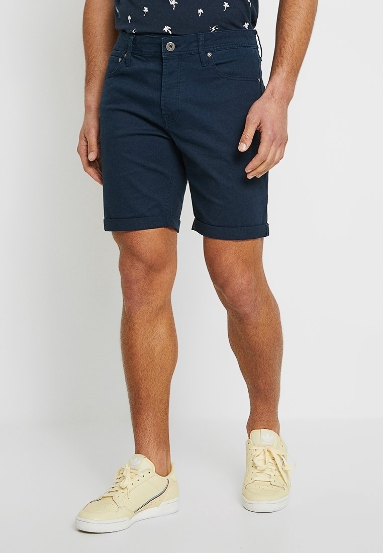 Shorts en coton bleu marine avec des revers, design à cinq poches et fermeture à bouton à l'avant. Portés avec des baskets jaunes.