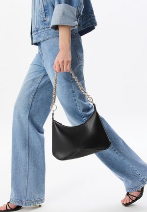 Bolso de hombro de cuero negro con base texturizada, forma curva y correa de cadena metálica, llevado por una persona con denim.
