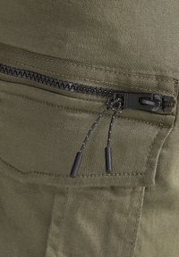 Nahaufnahme einer olivgrünen Stofftasche mit einem schwarzen Kunststoff-Reißverschluss und zwei schwarzen Kordelzügen an einem Kleidungsstück.