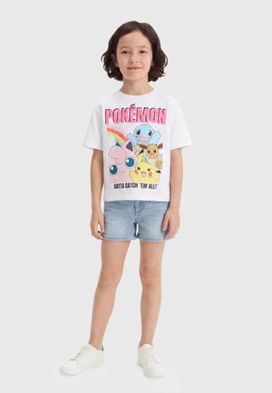 Enfant aux cheveux foncés mi-longs portant un t-shirt blanc Pokémon avec des personnages et un short en jean bleu clair, debout avec des baskets blanches.