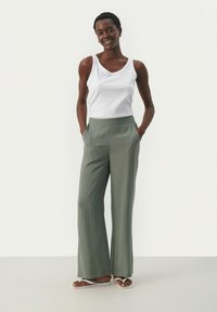 Pantalon léger taille haute, vert olive, avec des jambes larges et des poches latérales, associé à un débardeur blanc et des tongs.
