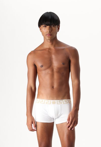 White boxer briefs with a gold Greek key pattern on the waistband, made from a soft fabric. Model has a short, rounded hairstyle.
