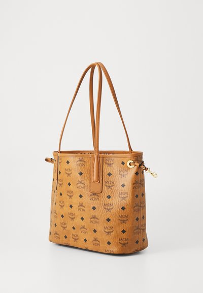 Tan lederen tote bag met zwart MCM-logo patroon, dubbele schouderbanden en gouden sluitingdetail op een effen achtergrond.