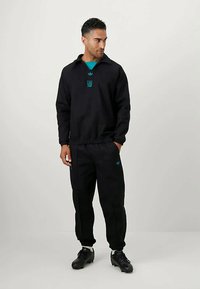 Melns sporta treniņtērps ar ceturtdaļas rāvējslēdzēja augšu. Iezīmējas ar tirkīza akcentiem un zīmolu. Biksēm ir elastīgi aproces un diskretais logotipa dizains.