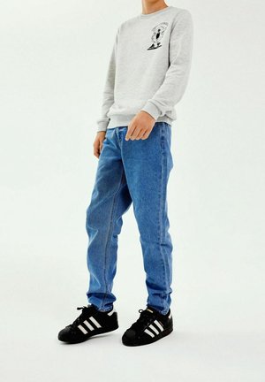 Sudadera gris con un gráfico negro, jeans de mezclilla azul claro y zapatillas negras con rayas blancas. Atuendo casual, diseño sencillo.