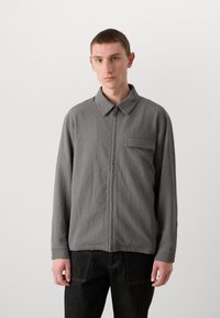 Chaqueta gris con cremallera, fabricada con un tejido texturizado, con cuello tipo polo, un bolsillo en el pecho y corte relajado. Se lleva con pantalones oscuros.
