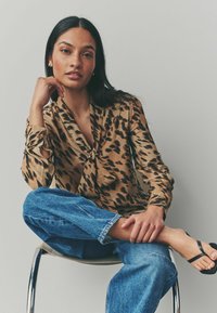 Blouse marron avec imprimé léopard noir, présentant un ourlet à nouer à l'avant, des manches longues et une texture lisse. Associée à un jean en denim bleu et des sandales.