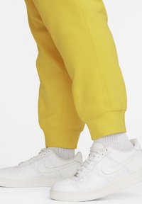 Pantalones de chándal amarillos con puños acanalados combinados con zapatillas Nike blancas y calcetines de textura gris claro. Las zapatillas tienen un acabado de cuero suave.