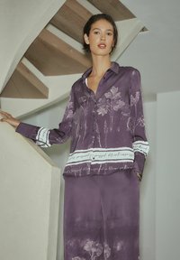 Lilla silkesbluse med blomstermønster og stribede detaljer. Har krave, knaplukning og lange ærmer. Glat tekstur og afslappet pasform.