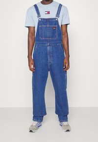 Modré denimové kalhotové lacláče s nastavitelnými ramínky, přední kapsou a volným střihem. Kombinované s světle modrým tričkem a sportovními teniskami.