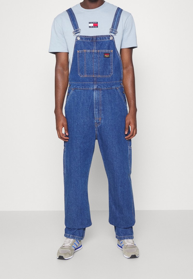 Modré denimové kalhotové lacláče s nastavitelnými ramínky, přední kapsou a volným střihem. Kombinované s světle modrým tričkem a sportovními teniskami.