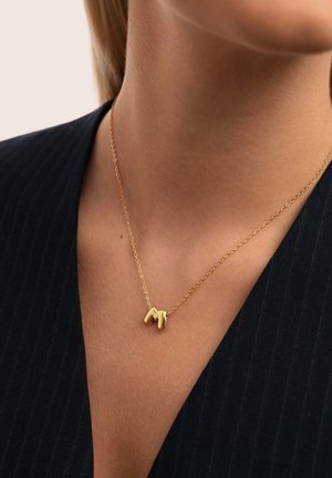 Collar de cadena dorada con un pequeño colgante abstracto y brillante de oro, llevado por una persona de piel clara y una blusa negra con rayas finas.