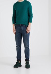 Man som bär en teal långärmad tröja, mörkblå upprullade jeans och gröna och svarta sneakers mot en plain vit bakgrund.