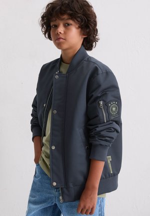 Dark navy bomber jacket with zippers, snap buttons, and a patch on the sleeve. Features ribbed cuffs and a smooth texture. Worn over a green shirt.